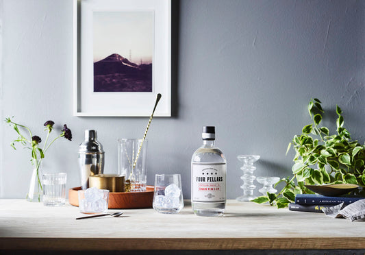 A bottle of Four Pillars Gin Cousin Vera Gin sitting on a table with barware, a glass full of ice and some plants.