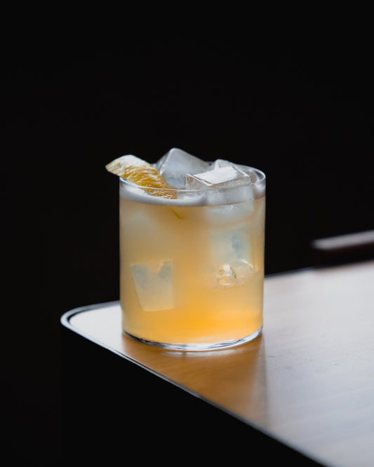 A non-alcohlolic yellow cocktail in a rocks glass with ice and a lemon twist on a wooden surface.