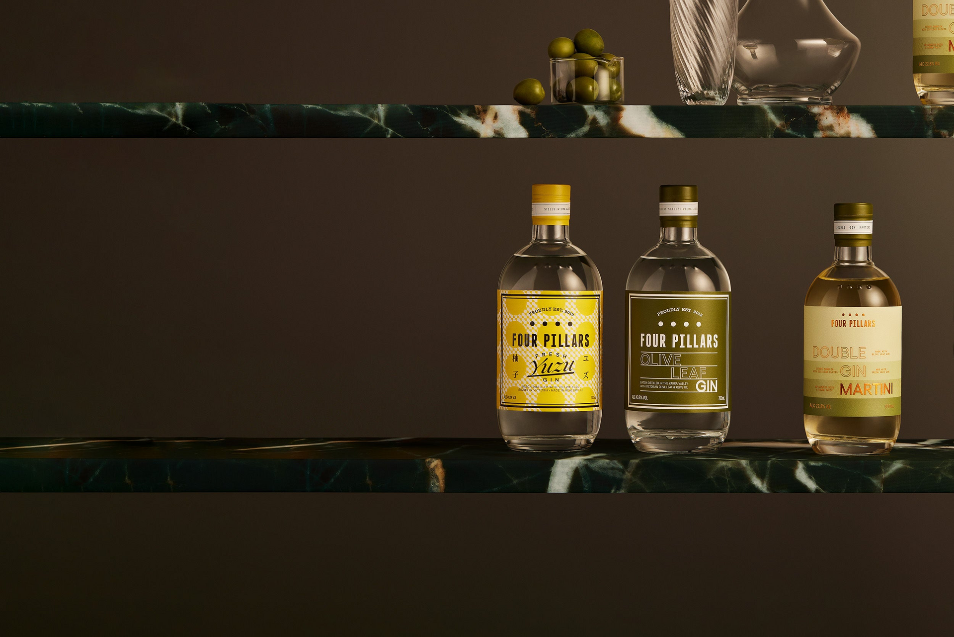 Two marble shelves with 3 bottles of Four Pillars Gin sitting on one, with olives and glassware on another. The three gins are Fresh Yuzu Gin, Olive Leaf Gin and the bottled cocktail Double Gin Martini.