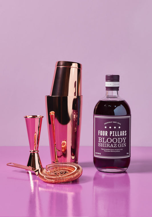 Four Pillars Shake It Bloody Better Bundle featuring a bottle of Bloody Shiraz Gin alongside a rose gold cocktail shaker, strainer, jigger, and bar spoon, all set against a vibrant purple background.
