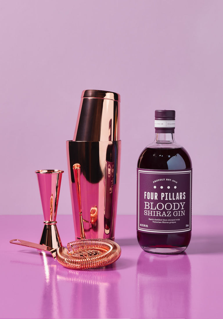 Four Pillars Shake It Bloody Better Bundle featuring a bottle of Bloody Shiraz Gin alongside a rose gold cocktail shaker, strainer, jigger, and bar spoon, all set against a vibrant purple background.