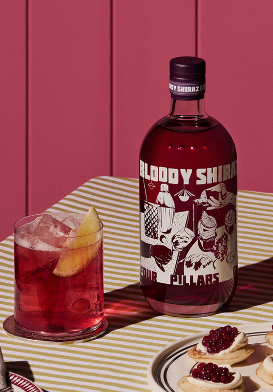 A Four Pillars Gin bottle of their 2024 limited edition Bloody Shiraz Gin, with label art by Kris Leombruni, next to a bloody shiraz g&T with an orange garnish, on a table with a striped table cloth, in front of a red wall.