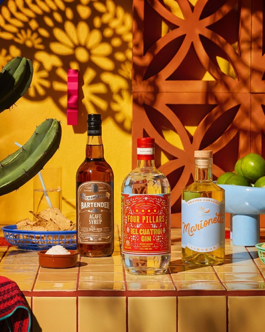 Three bottles of Mexican-style cocktails displayed on a tiled counter: a bottle of Bartender Agave Syrup, a bottle of Four Pillars Tequila Cucumber Gin, and a bottle of Marienette Tequila. The scene is styled with a cactus, lime, and a decorative wooden screen casting shadows on a yellow wall.