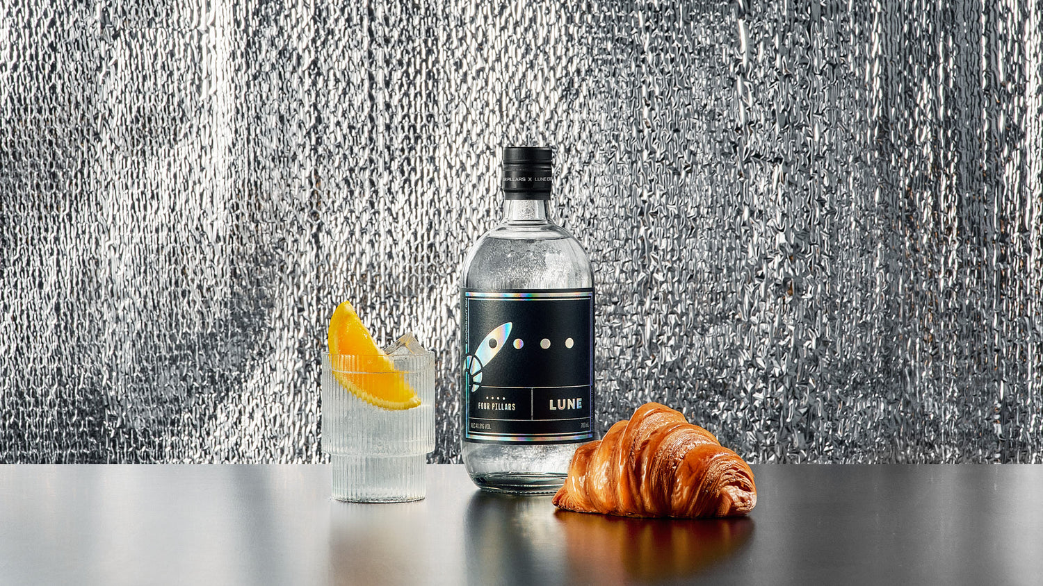 A bottle of Four Pillars X Lune Croissanterie collaboration gin, Croissant Gin, next to a traditional croissant and a gin and tonic garnished with a wedge of orange.