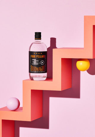 A bottle of Four Pillars Bandwagon Dry Gin sits on a peach-colored zigzag shelf with a yellow and a white ball beside it against a pink background.