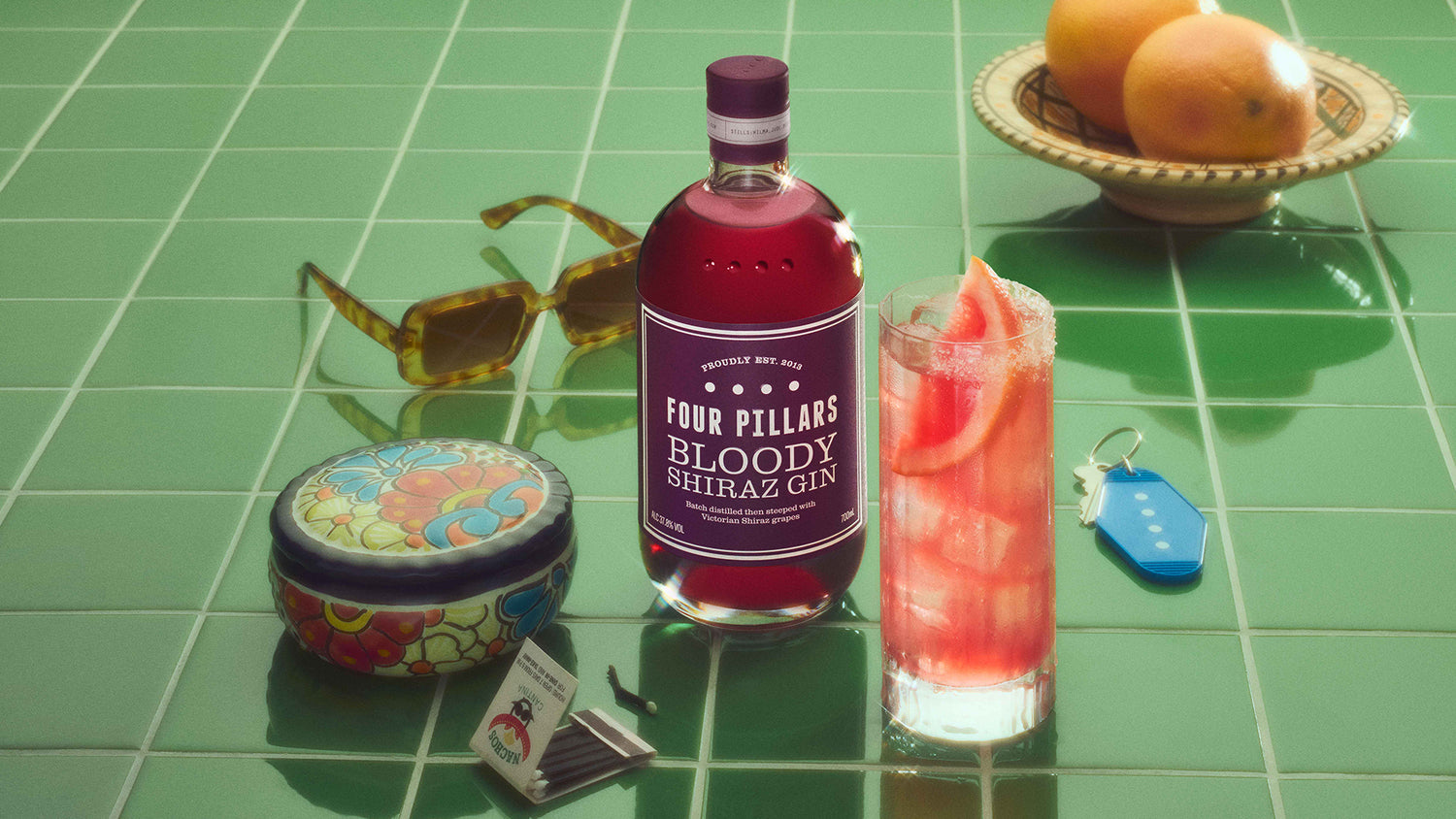 A bottle of Four Pillars Bloody Shiraz Gin next to a tall glass containing a Bloody Paloma Ggn cocktail, garnished with a slice of grapefruit.