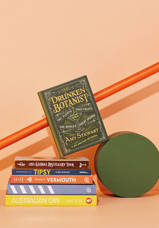 A pile of books on gin and various gin cocktails in front of an orange wall.