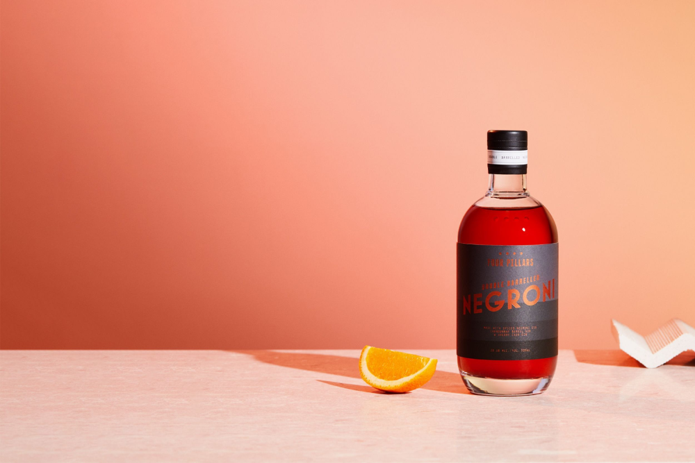 Double barrel Negroni, a bottled cocktail by Four Pillars gin with a sleek black label and a red liquid, sitting on a pink table next to a slice of orange.