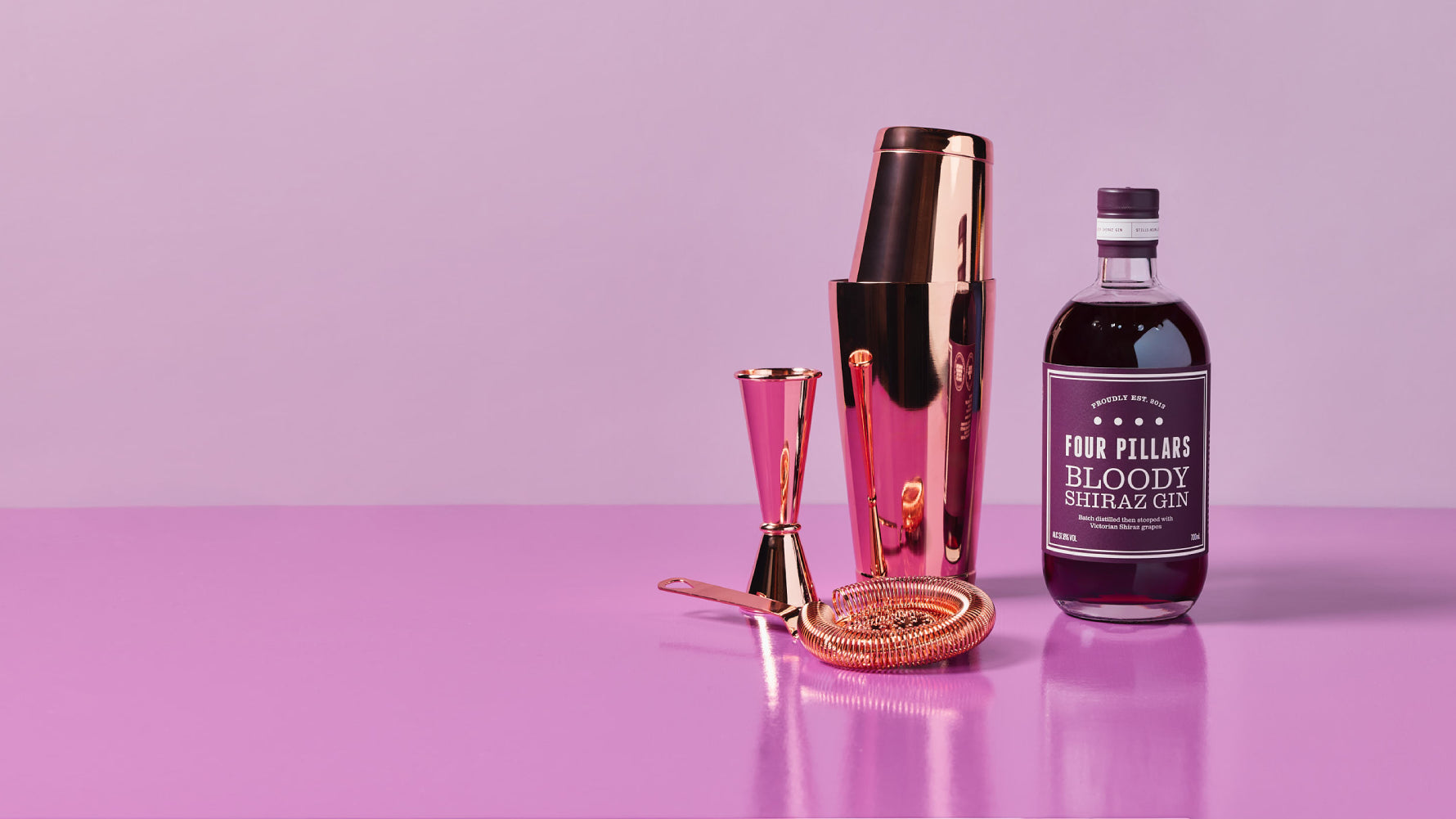 Four Pillars Shake It Bloody Better Bundle featuring a bottle of Bloody Shiraz Gin alongside a rose gold cocktail shaker, strainer, jigger, and bar spoon, all set against a vibrant purple background.