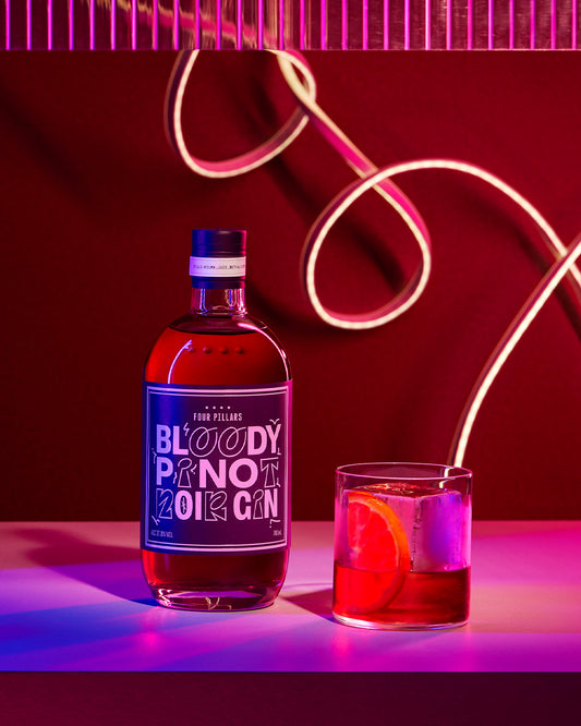 A beautifully lit bottle of Four Pillars Gin Bloody Pinot Noir gin next to a brightly lit gin cocktail in a short glass. 