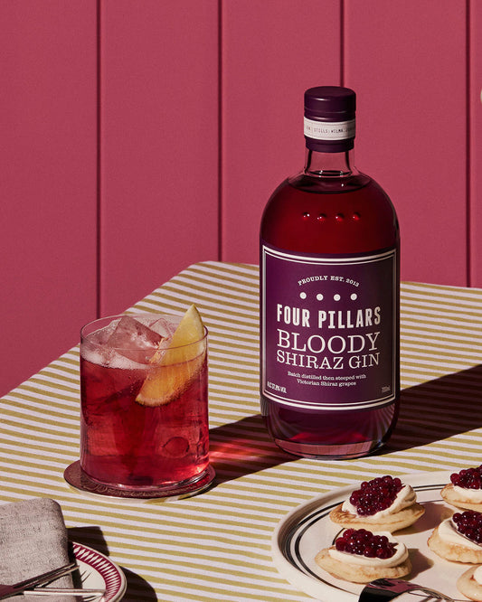 Four Pillars Gin Bloody Shiraz Gin bottle besides a bloody gin and tonic garnished with an orange. 
