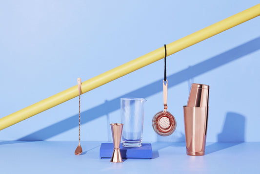 A selection of brass barware and cocktail mixing utensils in front of a blue wall. 