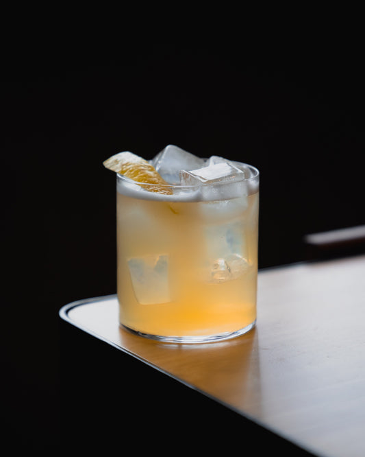 A non-alcohlolic yellow cocktail in a rocks glass with ice and a lemon twist on a wooden surface.
