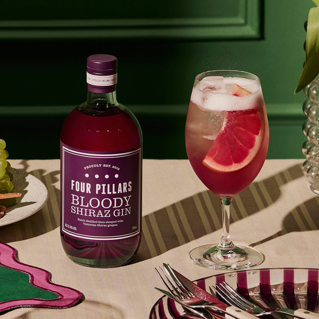 A dining table spread features a bottle of Four Pillars Bloody Shhiraz Gin next to a wine glass containing a Bloody Shiraz Gin Spritz cocktail. The cocktail is garnished with a slice of grapefruit.