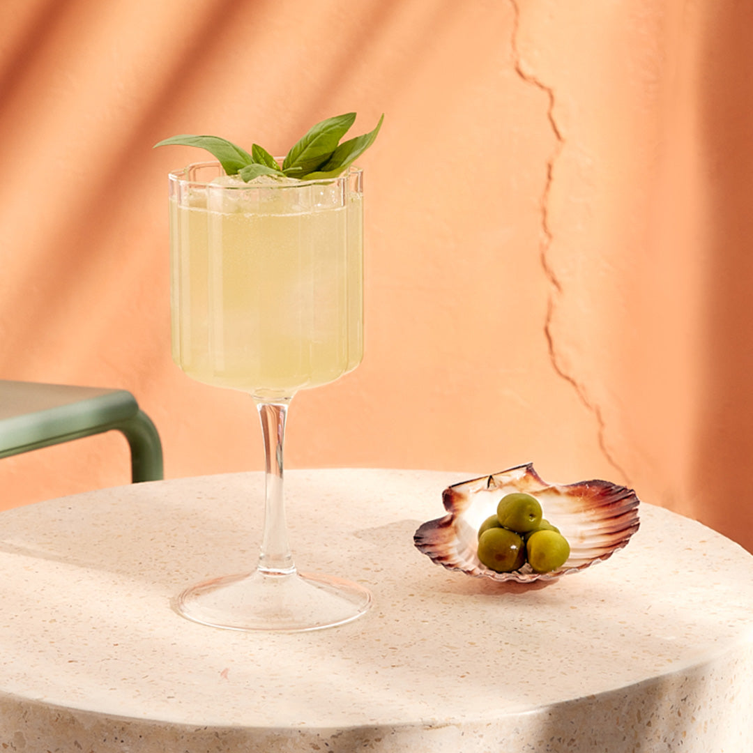A cocktail glass containing a Four Pillars Rare Dry Gin Spritz sits on a white stone block next to an open shell filled with green olives.