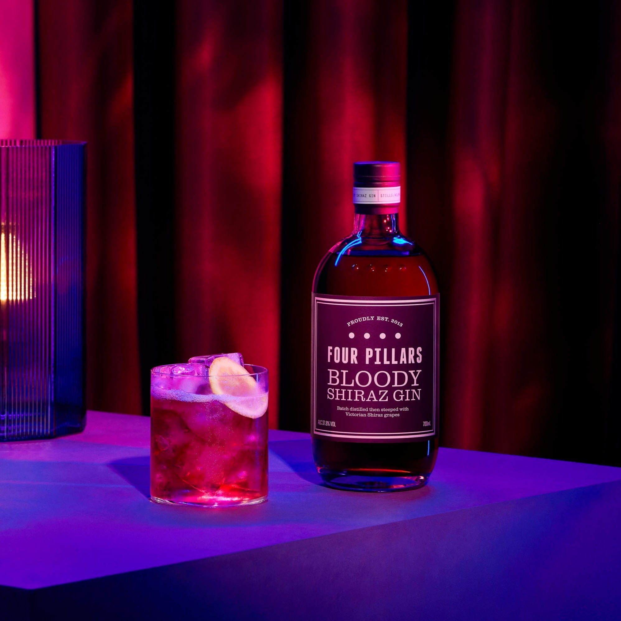 Bottle of Four Pillars Bloody Shiraz Gin with a G&T on a purple surface with a pink curtain background