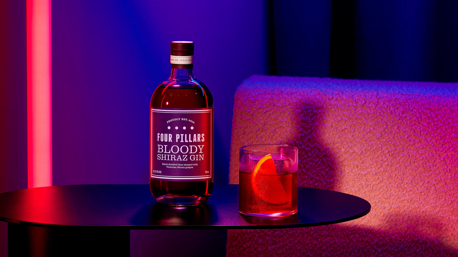 A Four Pillars Gin bottle of Bloody Shiraz Gin in a blue and red lit bar, on a table beside a cafe negroni cocktail garnished with an orange wedge. 