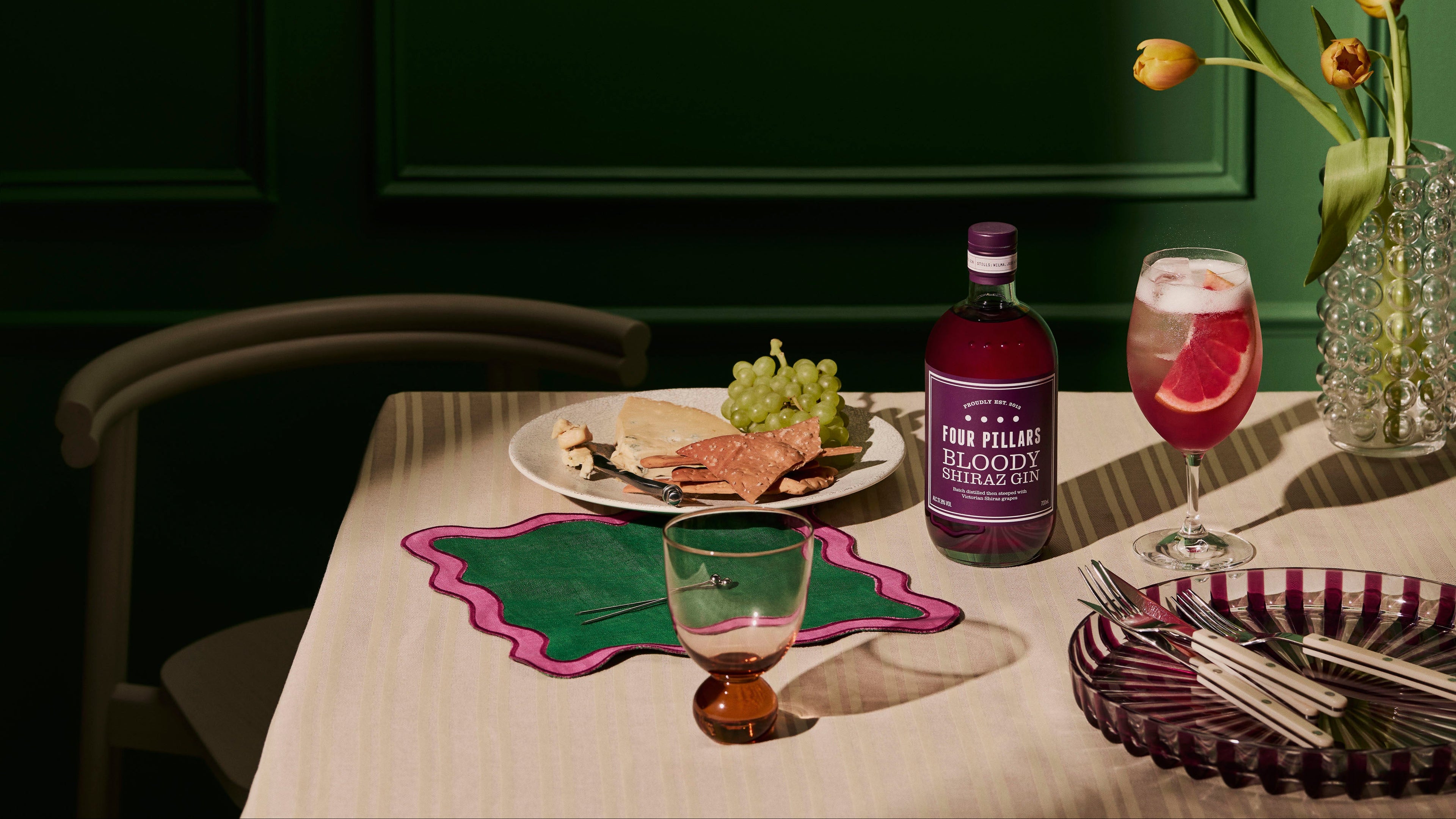 Dining table setup with a bottle of Four Pillars Gin, a glass of pink cocktail, and a plate of food.