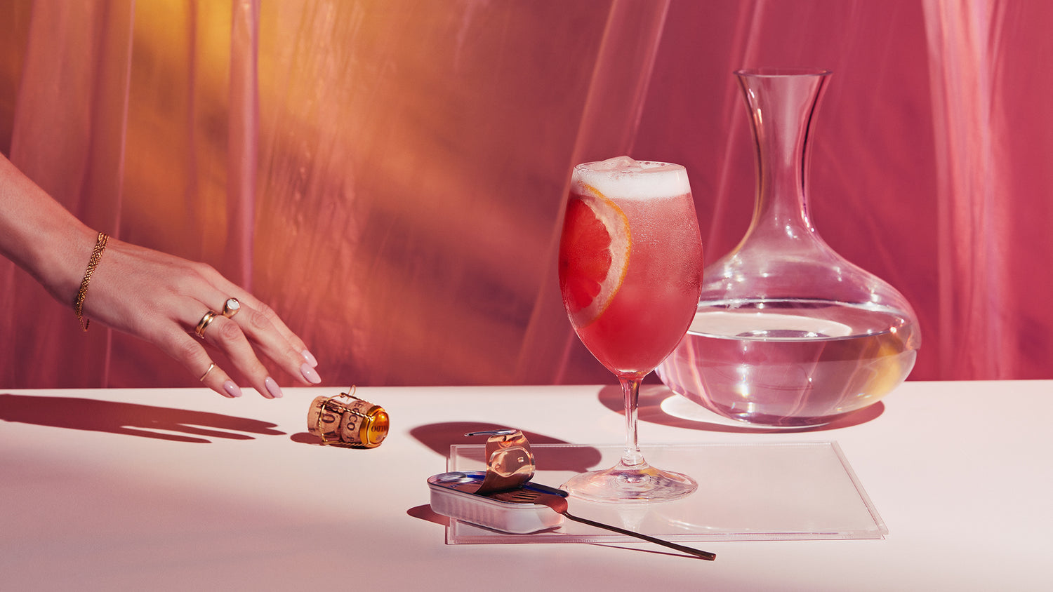 A hand wearing gold jewellery reaches for a large wine glass filled with a pink Gin Spritz. The Spritz is made with Four Pillars Bloody Shiraz Gin and grapefruit.