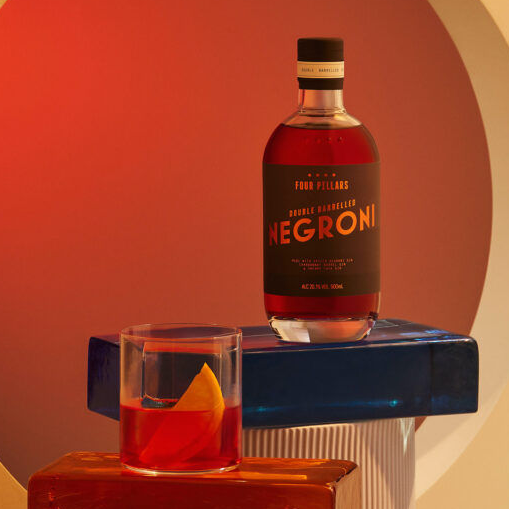 Bottle of Four Pillars Double Barrelled Negroni with a glass of Negroni on a blue stand against a warm gradient background.