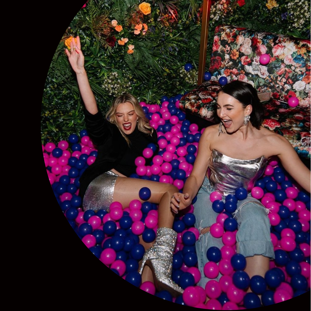 Two people in a ball pit with colorful balloons and text about Ballers Melbourne event.