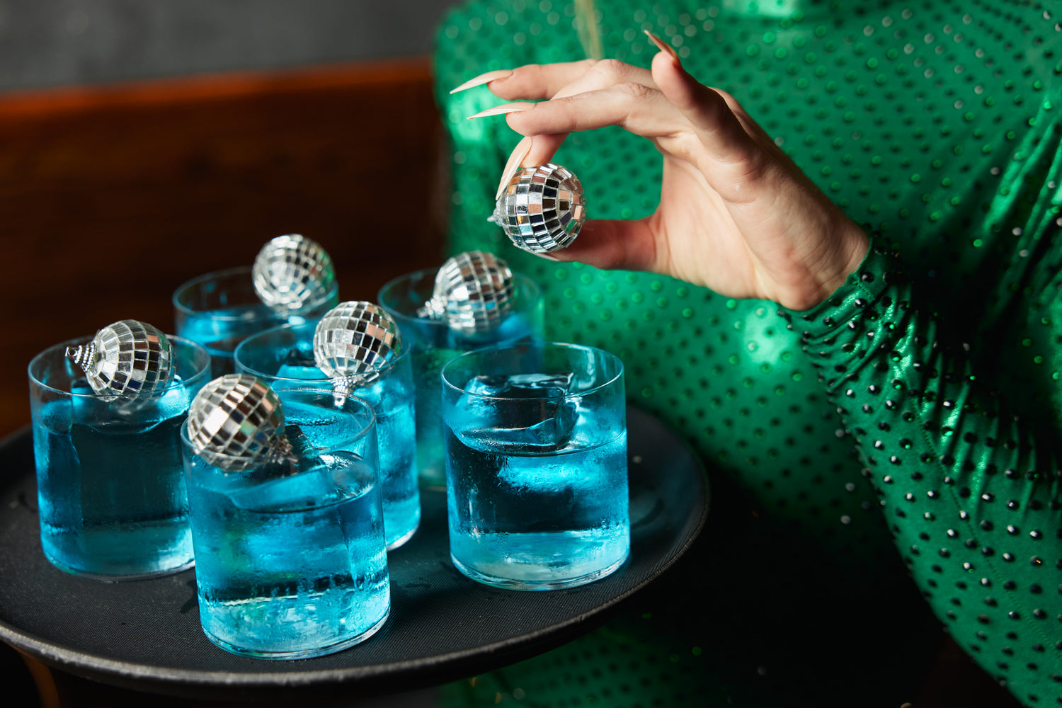 A drag queen holds a tray of bright blue coloured cocktails garnished with mini disco balls.