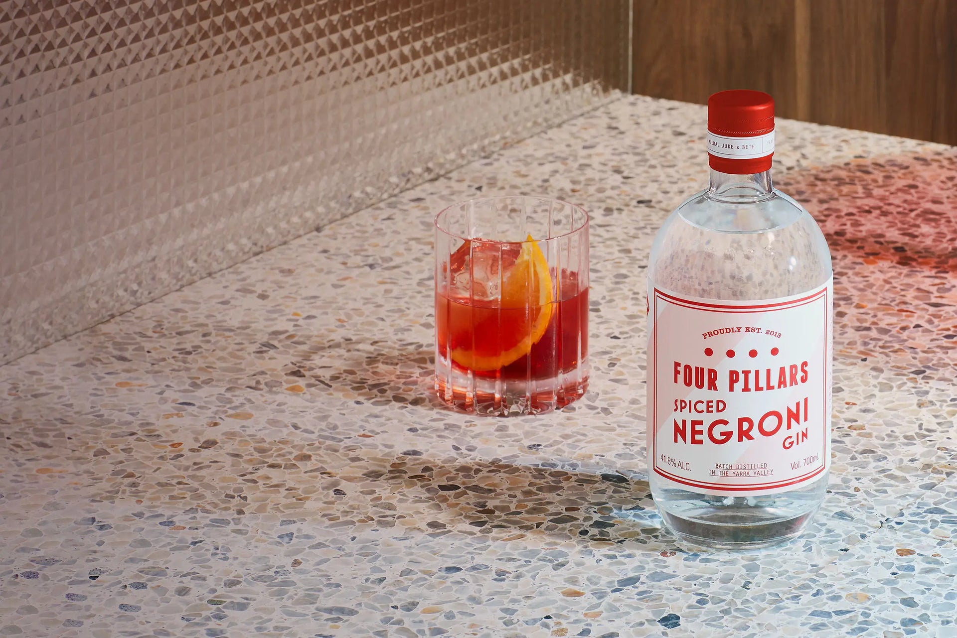 A bottle of Four Pillars Gin Spiced Negroni Gin sitting on a retro kitchen bench, next to a negroni with a garnish of orange. 