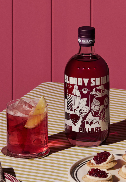 A Four Pillars Gin bottle of their 2024 limited edition Bloody Shiraz Gin, with label art by Kris Leombruni, next to a bloody shiraz g&T with an orange garnish, on a table with a striped table cloth, in front of a red wall. 