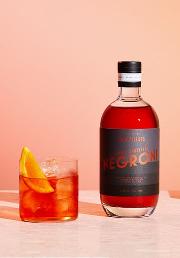 A bottle of Four Pillars Gin's ready-to-drink Double Barrelled Negroni cocktail sits beside a freshly poured Negroni, garnished with an orange slice, all set against a vibrant pink background.