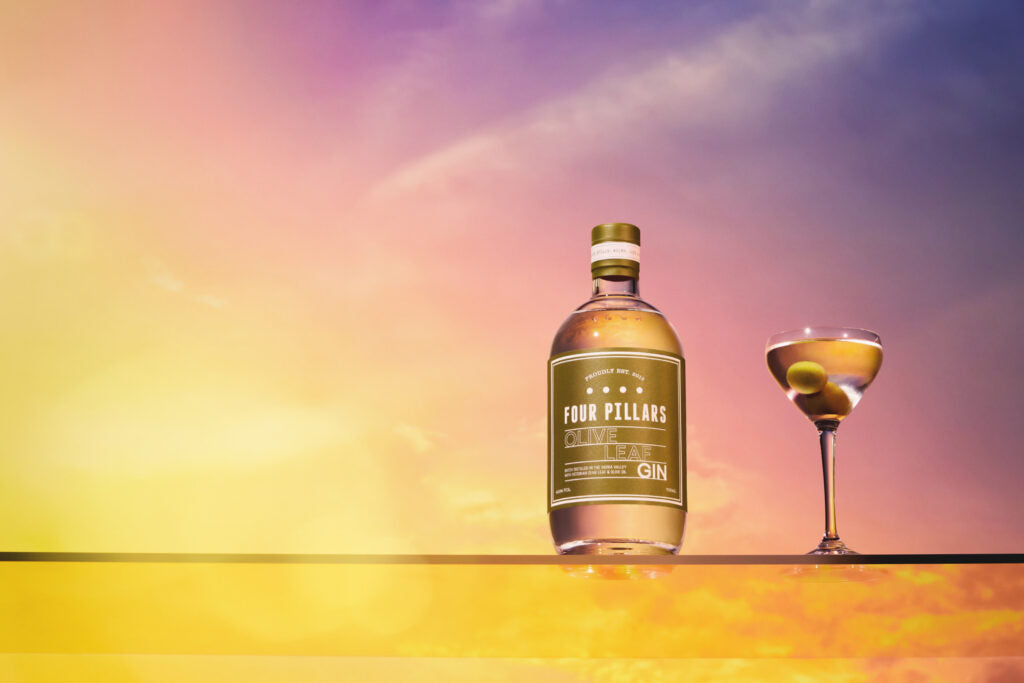 A bottle of Four Pillar's Gin Olive Leaf Gin next to a dirty martini with olives in front of a purple, blue and yellow sky.