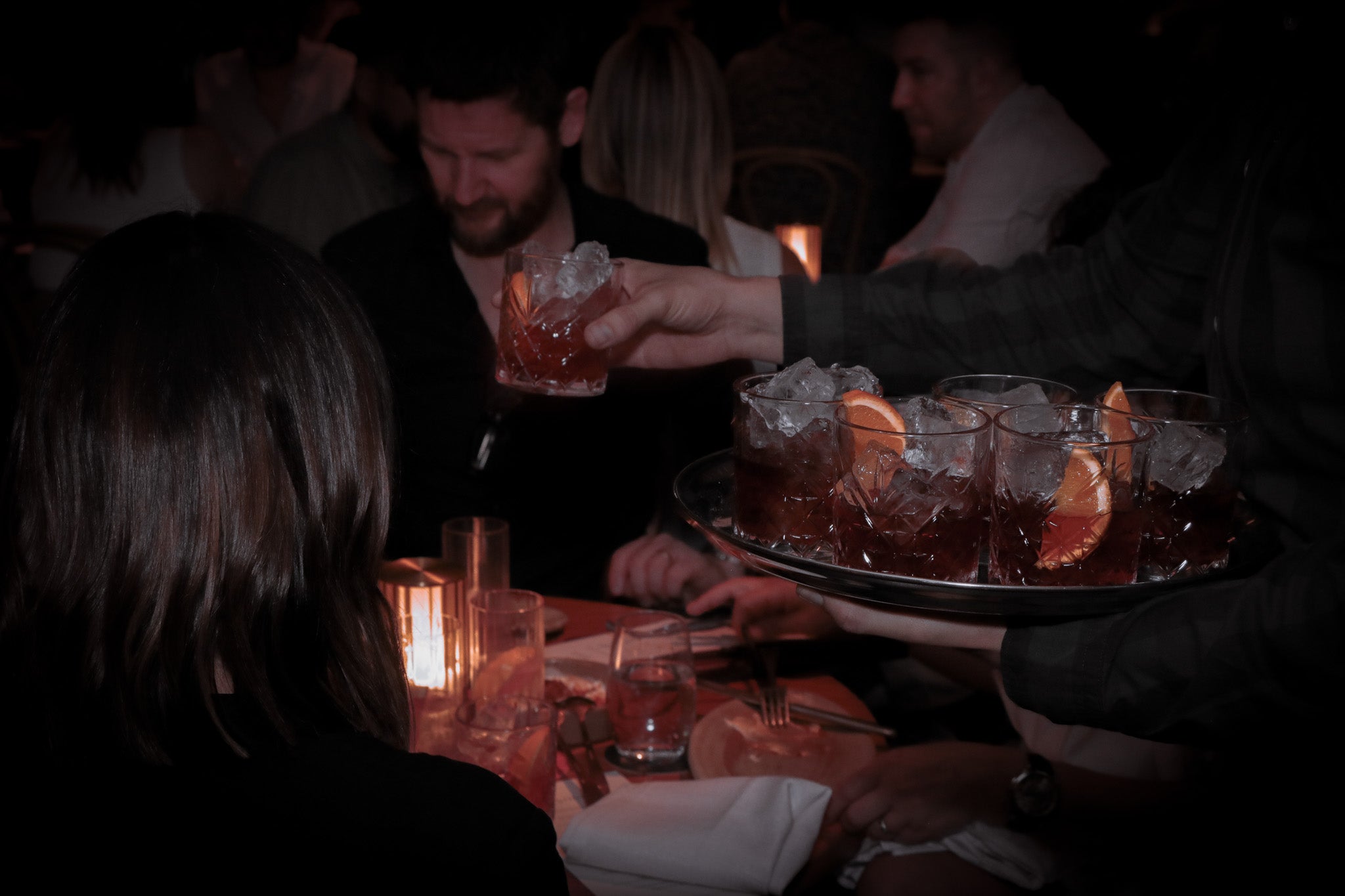 A tray of Negronis is handed to guests sitting at a dining table.