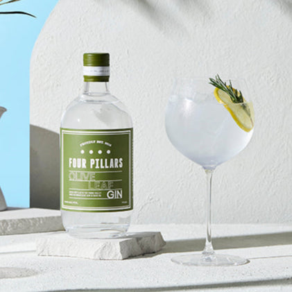 Four Pillars Olive Leaf Gin bottle with a glass of gin and tonic on a stone surface with a blue sky background