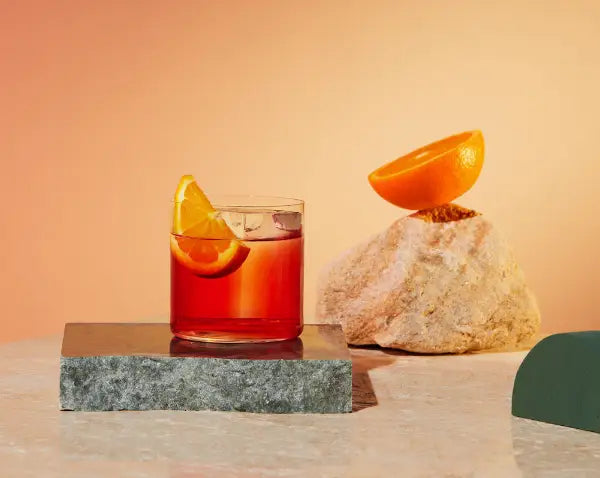 A short glass filled with a negroni cocktail and a wedge of orange, sitting on a slab of marble beside half an Orange sitting on a rock. 