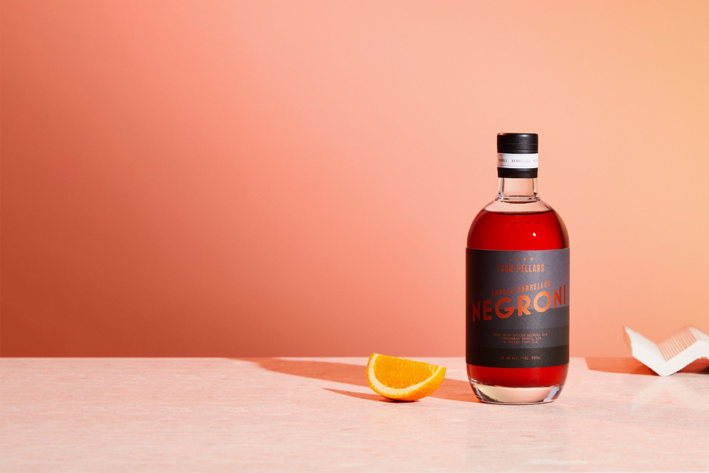 Double barrel Negroni, a bottled cocktail by Four Pillars gin with a sleek black label and a red liquid, sitting on a pink table next to a slice of orange. 