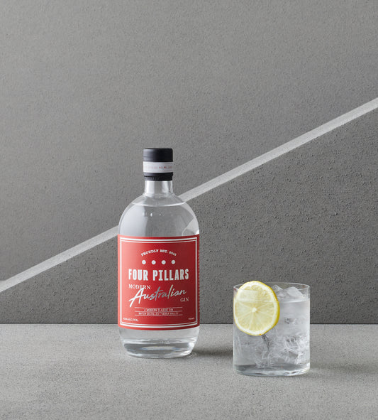 A bottle of Four Pillars Gin, Modern Australian gin, with it's bright red label sitting in front of a grey back ground and next to a refreshing gin and tonic with a lemon garnish.