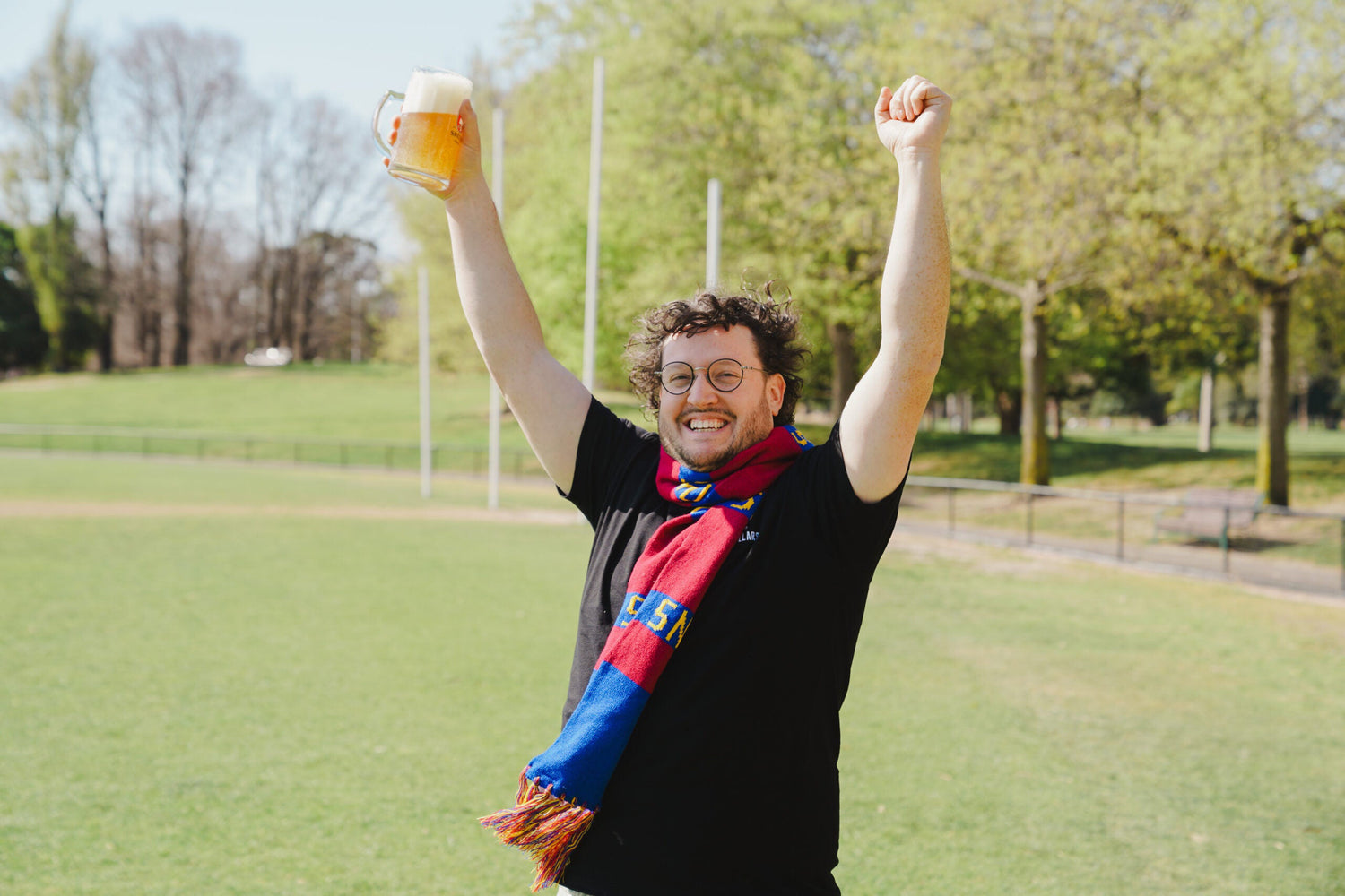 Nick Tesar wearing a colourful, Melbourne AFL team scarf, holding a glass of beer, and raising both arms in celebration, stands on a grassy field at a sunny sporting event.