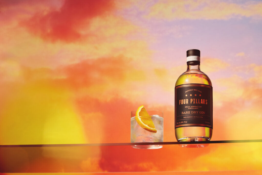 A gin and tonic with an orange garnish next to a bottle of Four Pillars Gin Rare Dry Gin in front of a vibrant orange sky.