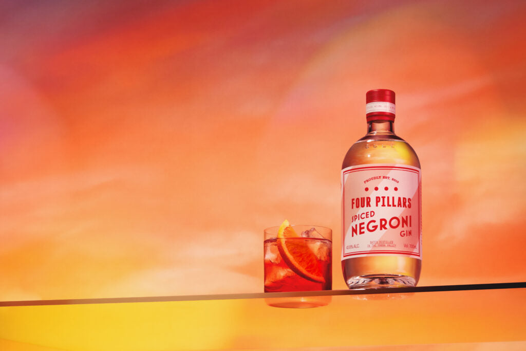 A bottle of Four Pillars Gin Spiced Negroni Gin next to a delicious red negroni cocktail in front of a bright red sky.