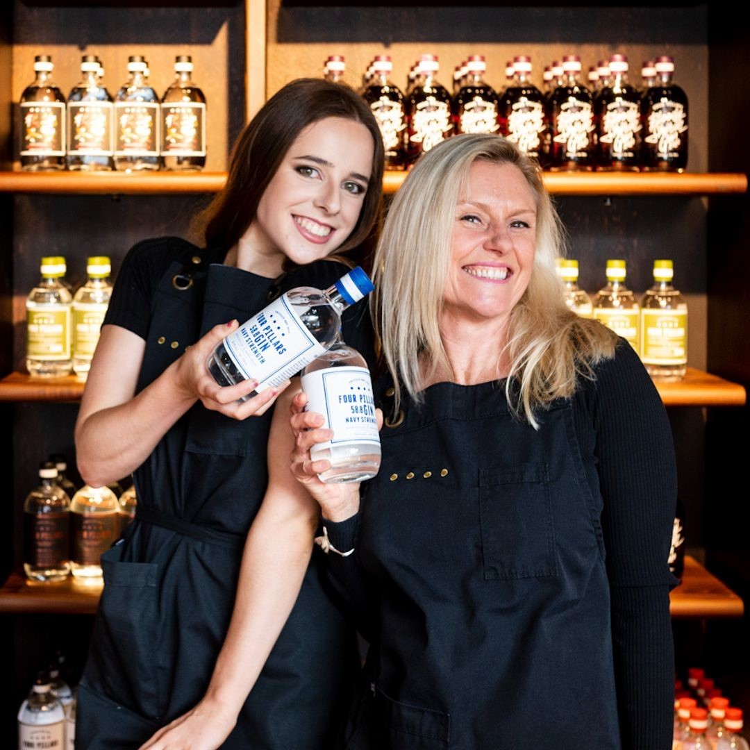 Two women holding a bottle of Four Pillars Navy Strength Gin, standing in front of shelves with more bottles.