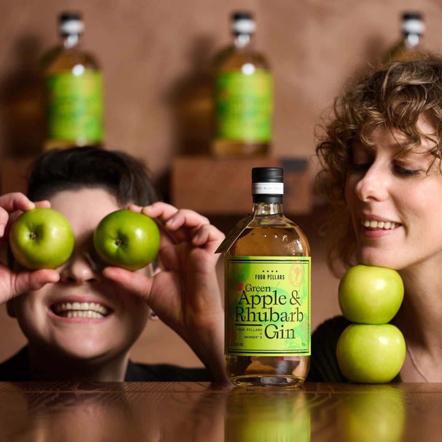 Two people with green apples and a bottle of Four Pillars Green Apple & Rhubarb Gin on a wooden surface.